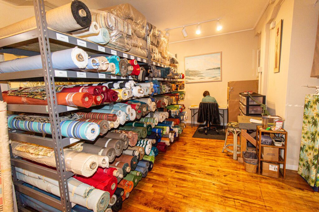 Dozens of large rolls of fabrics on shelves, with a person sitting at a desk in the background to the right of the fabric shelving.